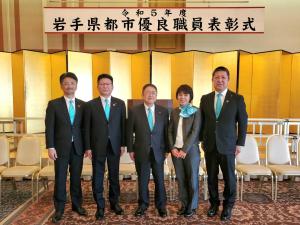 「令和5年度岩手県都市優良職員表彰式」の横断幕と金屏風、白い椅子がたくさん並べられた室内で水色のネクタイをした男性4名と水色のスカーフをした女性1名が並んで記念撮影をしている写真