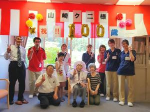 窓際から吊るされた紅白幕に「祝100歳」と書かれた装飾が施された部屋で、椅子に座る満100歳を迎える山崎クニさんを中心に、市長やご家族、施設の関係者達が写っている写真