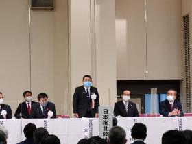 全国大会の会場で白のリボン胸章をつけた市長が自席で立って、参加者の方々にあいさつをしている写真