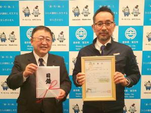 バックパネルの前で目録を持つ市長と額縁を持つ眼鏡をかけた男性が写っている写真