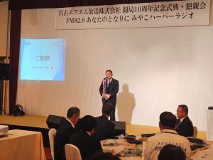 横断幕と大きなスクリーン、ステージ中央にマイクが設置され、マイクの前に立つ市長が挨拶を行っている記念式典の様子の写真