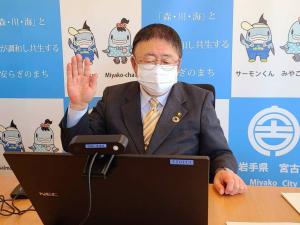 椅子に座りパソコンの画面を見ながら右手を顔の横に掲げている市長の写真