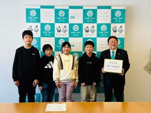宮古市のバックパネルをバックに花輪小児童の学生4名が並び、能登半島地震災害義援金の箱を市長が持って記念撮影をしている写真
