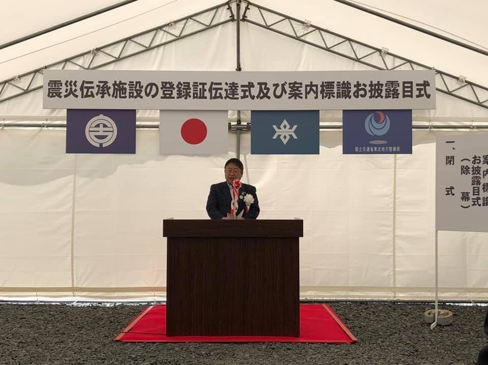 白いテント下の会場に「震災伝承施設の登録証伝達式及び案内標識お披露目式」と書かれた横断幕と4つの旗が掲げられ、檀上の前で挨拶を行っている市長の写真