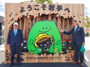 森をかたどった緑のキャラクターが「わんこきょうだい」を笑顔で包み込むシンボルマークを中央に「ようこそ岩手へ 第73回全国植樹祭いわて2023」と書かれた杉の木で作られたウェルカムボードの前で、キャップを被った市長と、左手に緑色のキャップを持ち右手をウェルカムボードに向けている副市長の写真