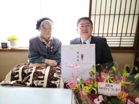 お祝いの花が置かれたご自宅の居間で、市長が市からの記念品を持ち、三野宮ミヱさんと撮影した写真