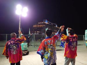 夜に出港するレガッタにライトで照らされた港から手を振る、法被を着た市長ら3人の男性の後ろ姿の写真