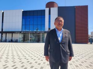 茶色や白色、グレー色の外壁とガラス張りで施され、中央のエレベーター部分が円柱に造られている複合施設「えみらん」の建物前に立つ市長の写真