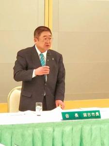 岩手県市長会議でコップが置かれ、「会長 宮古市長」の名札が置かれた机で市長が立ちながら挨拶をしている様子を写した写真