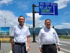 道路上に設置された青い道路標識を背に横に並ぶ市長と関係者の男性の写真