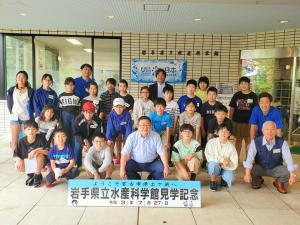 足元に置かれた「岩手県立水産科学館見学記念」と書かれたパネルの後方で市長を中心に多くの小学生たちが集まって写る記念写真