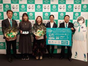 バックパネルの前で市長と女性がプロジェクトのボードを持ち、副市長と女性がフラワーアレンジメントを持ち、女性がアクセサリーを持っている写真