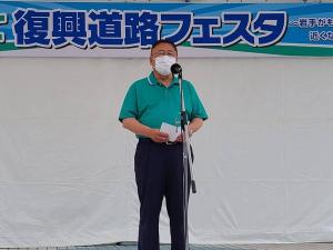 「復興道路フェスタ」と書かれた横断幕が掲げられた会場の前で、スタンドマイクの前に立ち話をする様子の市長の写真