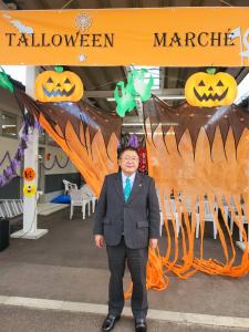 「TALLOWEEN MARCHE(タロウィーンマルシェ)」と書かれた横断幕やかぼちゃやオレンジ色の幕で飾り付けられた会場の前で立って写真に写る市長の写真