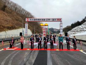 開通を祝う看板の下で割れたくす玉を背景に、道路に敷かれたレッドカーペット上で市長、鈴木俊一衆議院議員、階猛衆議院議員、高橋ひなこ文部科学副大臣、木戸口英司参議院議員、横沢高徳参議院議員でテープカットをしている写真