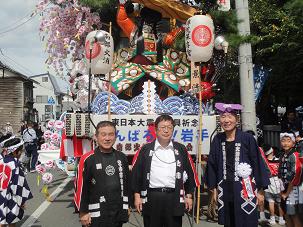 人形や造花等で創られた山車の前で、消防団のはっぴを着た市長と男性二人が映っている写真