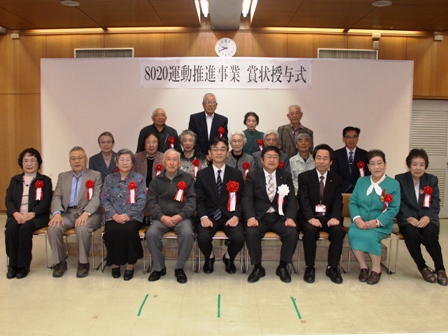 「8020運動推進事業賞状授与式」と書かれた横断幕の下、市長と関係者と、赤リボンを胸に付けた受賞者の男性や女性が横3列に並んでいる写真