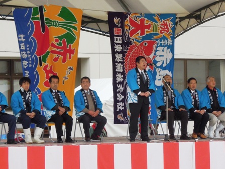 野外ステージに色鮮やかな青やオレンジの大漁旗がステージ中央後方に吊るされており、青色のはっぴを着た市長がステージ中央でマイクスタンドの前に立ち、その後ろにはっぴを着た関係者達が椅子に座っている写真