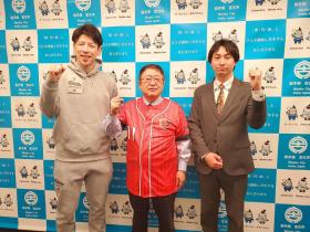 赤いユニフォームを着た市長をはさんで、岩手ビッグブルズの三浦崇代表取締役と千葉慎也選手がガッツポーズをしている写真