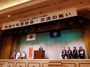 「在仙岩手県人会創立108周年 令和5年度総会・交流の集い」と書かれた横断幕が掲げられらステージの左右に胸章リボンをつけた方々が座り、中央に設置された檀上に男性が立ち話をしている様子の写真