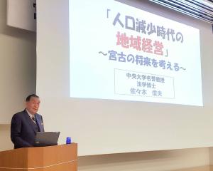 画面が映し出されたスクリーンの横の演台の前に立ち講演するスーツを着た男性の写真