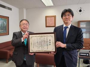 壁に絵画が飾られ応接用の椅子が設置された室内で、青山学院女子短期大学の河見誠学長と市長が一緒に額入りの感謝状を持って並んでいる写真