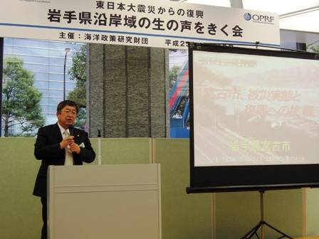 岩手沿岸の生の声をきく会と書かれた横断幕のあるステージで、山本市長がスクリーンに映し出された資料を用いて、宮古市の現状を報告している写真