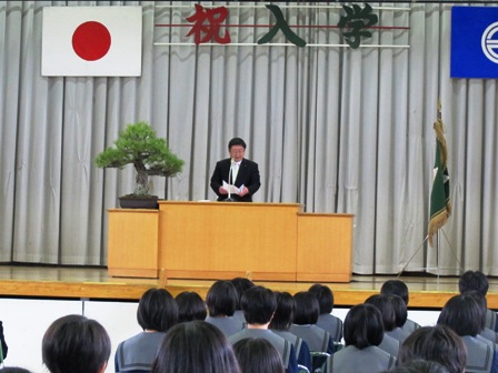 「祝 入学」と象られた文字がステージの上に飾られており、制服姿の生徒たちが整列する前で、演壇に立ち祝辞を述べる市長の写真