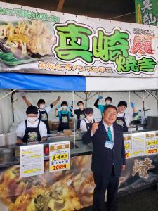 真崎焼きの屋台の前に立つ市長と、同じエプロンをした屋台の中で働く7名の方がガッツポーズをしている写真