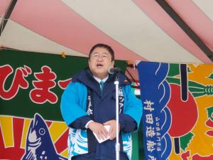 後方にカラフルな色の大漁旗が飾られたステージのマイクの前で、水色の法被を着た市長が挨拶を行っている写真
