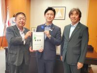 国旗が飾られた机の前で、市長を含む男性3名が並んで立ち、市長と男性で要望書を持っている写真