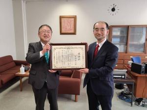 壁に絵画が飾られ本棚が設置された室内で、、吉岡光人様と市長が一緒に額入りの感謝状を持って並んでいる写真
