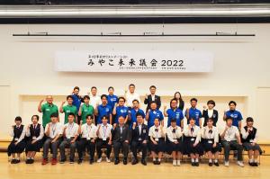 「みやこ未来議会2022」と書かれた横断幕が掲げられたステージ前列中央の椅子に市長と関係者の男性が座り、左右に参加した16名の高校生議員、後列にお揃いの青色や緑色のユニフォームを着た関係者の方々が3列に並んでいる集合写真