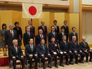 国旗が掲げられ金屏風が設置された会場で、佐藤1等陸佐と市長を中央に前列の椅子に7名座り、2列目と3列目に6名ずつ関係者が立っている集合写真