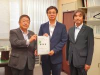 窓にブラインドがつけてある部屋で市長を含む男性3名が並んで立ち、市長と男性で要望書を持っている写真