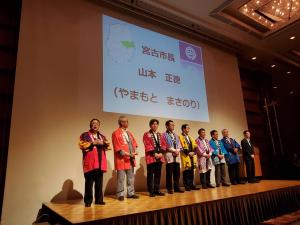 ステージ上にそれぞれ色の違う法被を着た8名の男性が並び、後方のスクリーンに「宮古市長 山本 正徳」と映し出されている写真