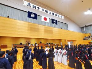 剣道着を着た学生たちが体育館で舞台の方を向き整列している写真