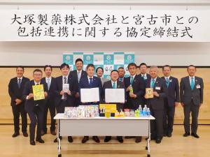「大塚製薬株式会社と宮古市との包括連携に関する協定締結式」と書かれた横断幕とバックパネルの前に長机の上に商品が並び、中央に締結書を持った市長と代表の男性が立ち、副市長と3名の関係者、後列に8名の関係者が並んでいる写真