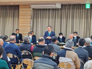 スーツ姿の関係者と参加者が向かい合って座り、中央に立って挨拶を行っている市長の写真