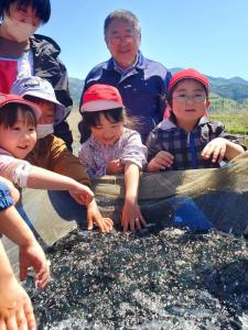 網に入った大量の鮭の稚魚を触ったり見つめている4人の児童とその後ろで笑顔を浮かべる市長の写真