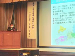 資料が映し出されたスクリーンの左側に「講演「震災復興の現状と今後」」のお題が書かれ、ステージ上の檀上で話をしている市長の写真