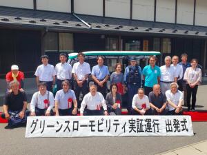 鍬ケ崎公民館前の屋外で「グリーンスローモビリティの実証運行出発式」と書かれた横断幕の後方に胸章リボンを付けた市長や関係者が2列に並んでいる記念写真