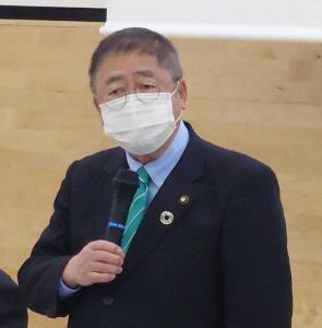 右手に持ったマイクを白いマスクを着けた口元に向け話している様子の市長の写真