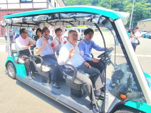 ドアがなく2名掛けで3列シートの明るいグリーン色のグリーンスローモビリティに乗車した市長や5名の関係者がカメラに向かって手を振っている試乗の様子の写真