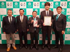 バックパネルの前で両手で目録を持った市長を中央に、右隣に額に入った賞状を持った男性、2名の関係者と1名の市の関係者が横一列に並んでいる記念写真