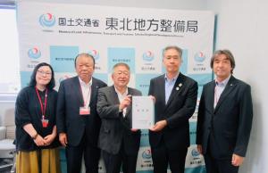 国土交通省東北地方整備局と書かれたバックパネルの前で、中央に東北地方整備局の山本巧局長と市長が一緒に要望書を持ち、3名の関係者が横一列に並んでいる写真