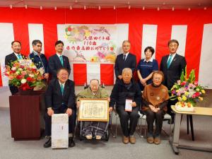 紅白幕を背景に、両脇を花瓶に生けられた花で囲まれた中で車いすに座って賞状を持つ久保田イチさんや、市長ら10名の集合写真