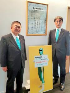 壁に飾られた額縁の前で、緑色の戦隊ヒーローが1名写った看板の両側に立つ市長とスーツを着た男性の写真