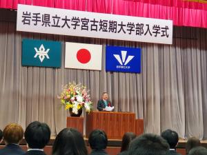 「岩手県立大学宮古短期大学部入学式」の幕、国旗、校旗が掲揚されたステージ上の演台で、市長が新入生に向けて祝辞を述べている写真
