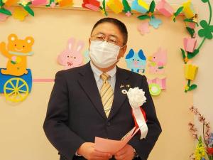 カラフルな色の折り紙で制作された花のアーチや乗り物に乗った動物たちが後方の壁に飾られた開所式の会場で、左胸に白色の胸章リボンを付けた市長が挨拶を行っている様子の写真